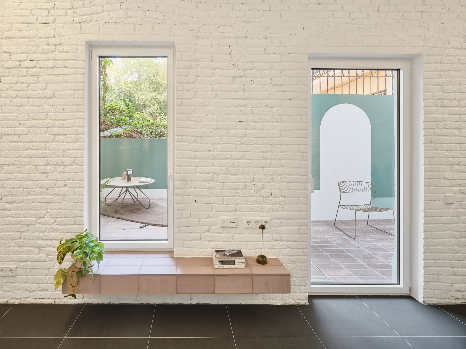 Interior view in CRÜ’s “La Dolors” renovation with two patio openings framed by whitewashed brick walls, a tiled bench, outdoor seating, and garden vegetation.